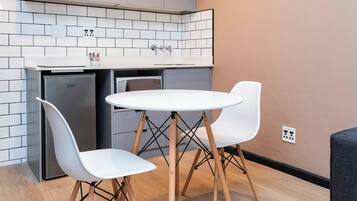 Eine kompakte Küche mit weiß gefliestem Fliesenspiegel, einem eingebauten Kühlschrank, einer Mikrowelle und einem kleinen Spülbecken. Im Hintergrund steht ein runder Tisch mit zwei weißen Stühlen und ein graues Sofa.