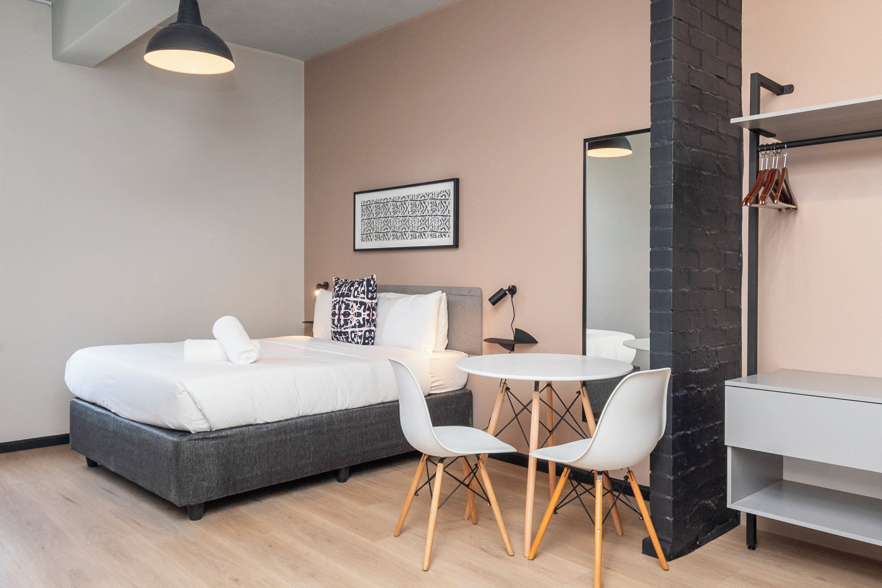 Ein modernes Hotelzimmer mit einem Bett, zwei weißen Stühlen, einem kleinen runden Tisch, einem Spiegel und einer Wand mit Ziegeloptik in Schwarz.
