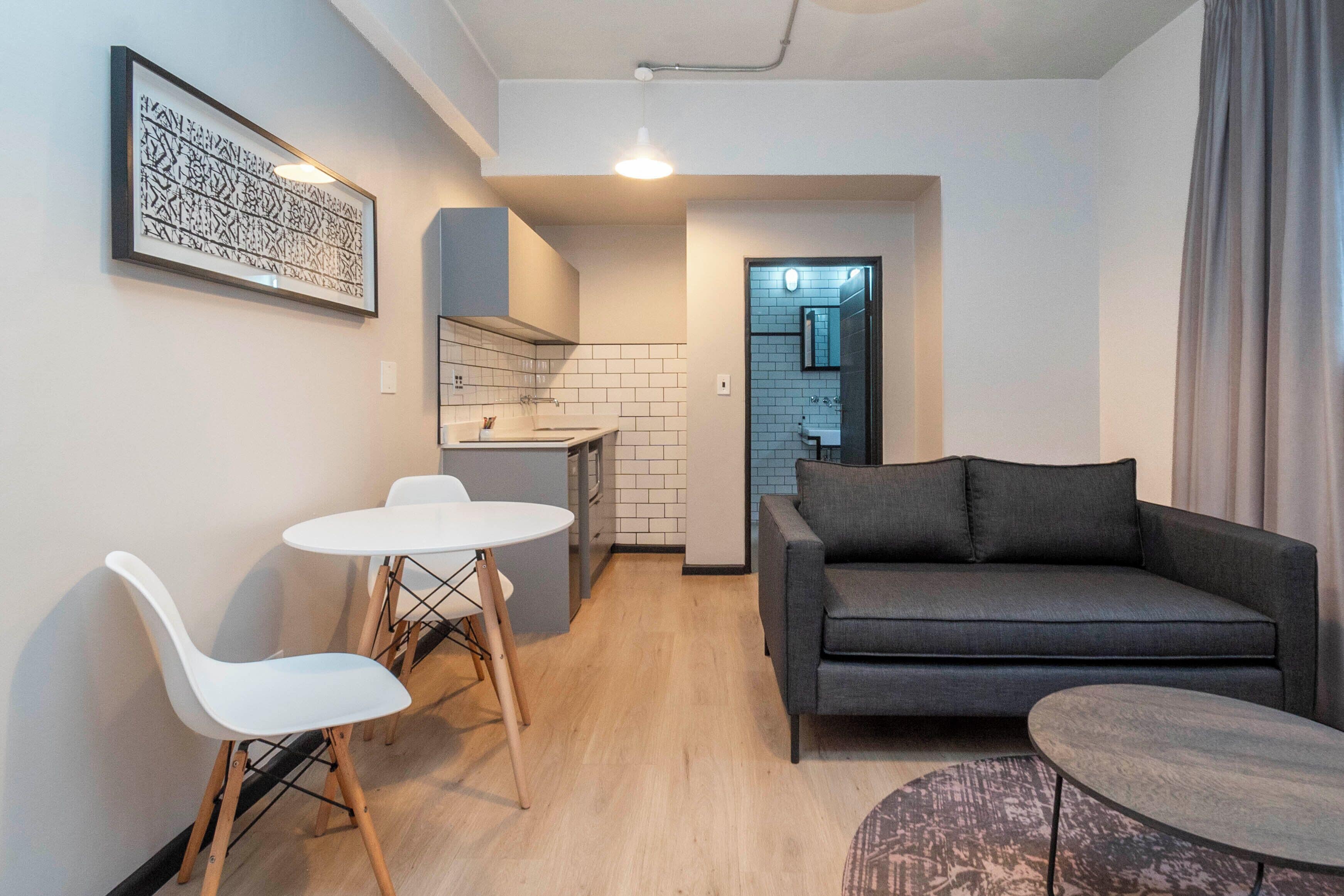 Un espacio habitable compacto con kitchenette, un sofá gris, una mesa de comedor con sillas y una pequeña mesa redonda.