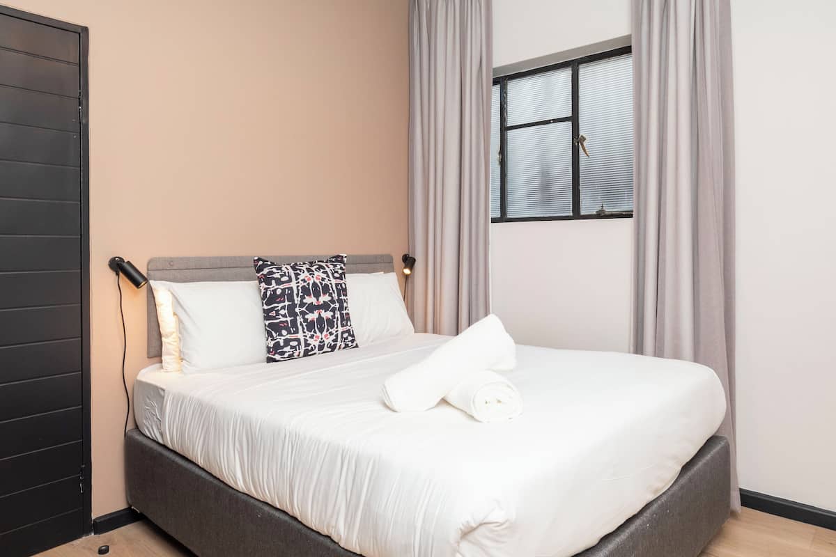Una cama bien hecha con una colcha blanca y almohadas, una lámpara de noche y una ventana con cortinas.
