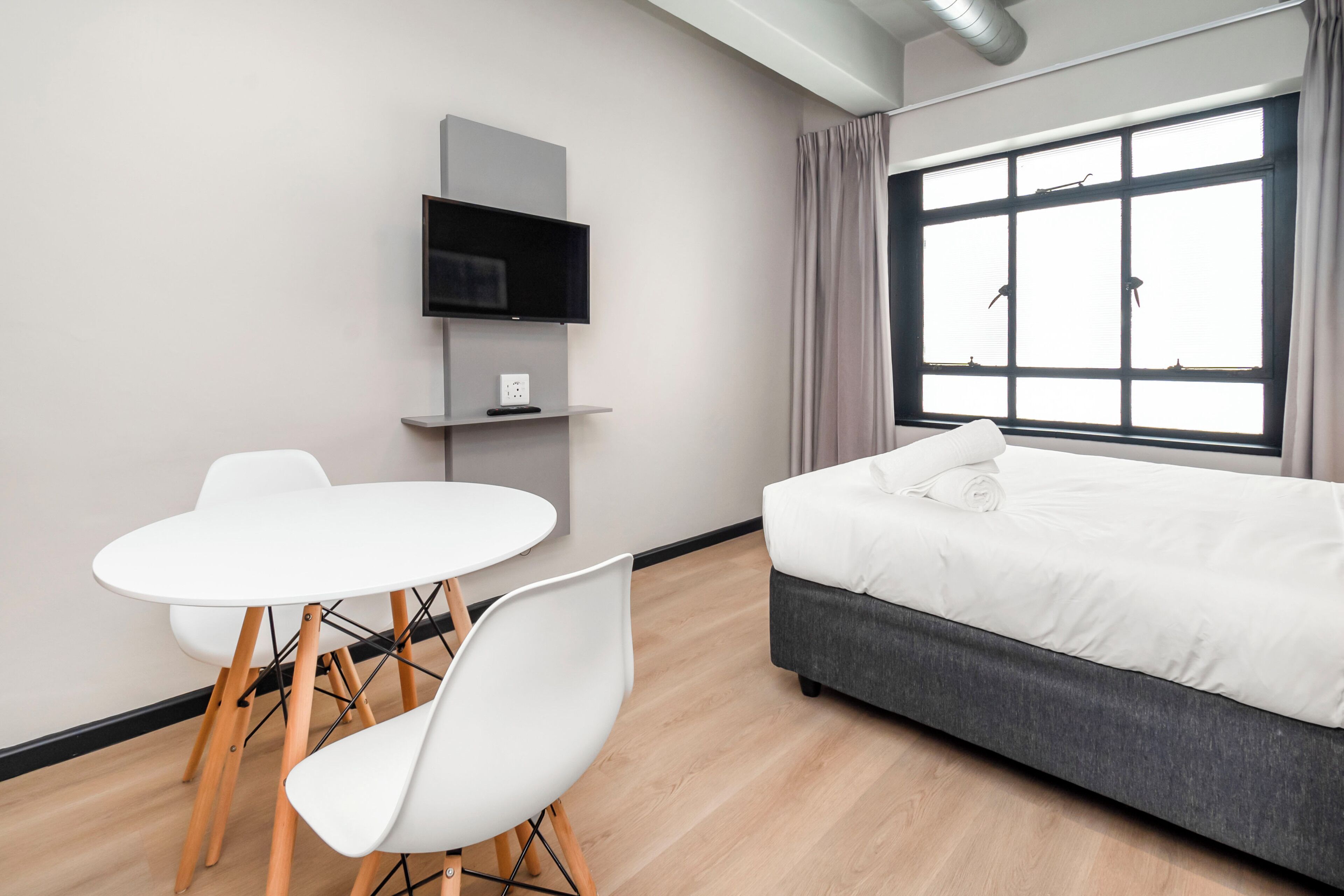 Quarto de hotel moderno com cama, televisão, mesa pequena com cadeiras e janela grande com cortinas.
