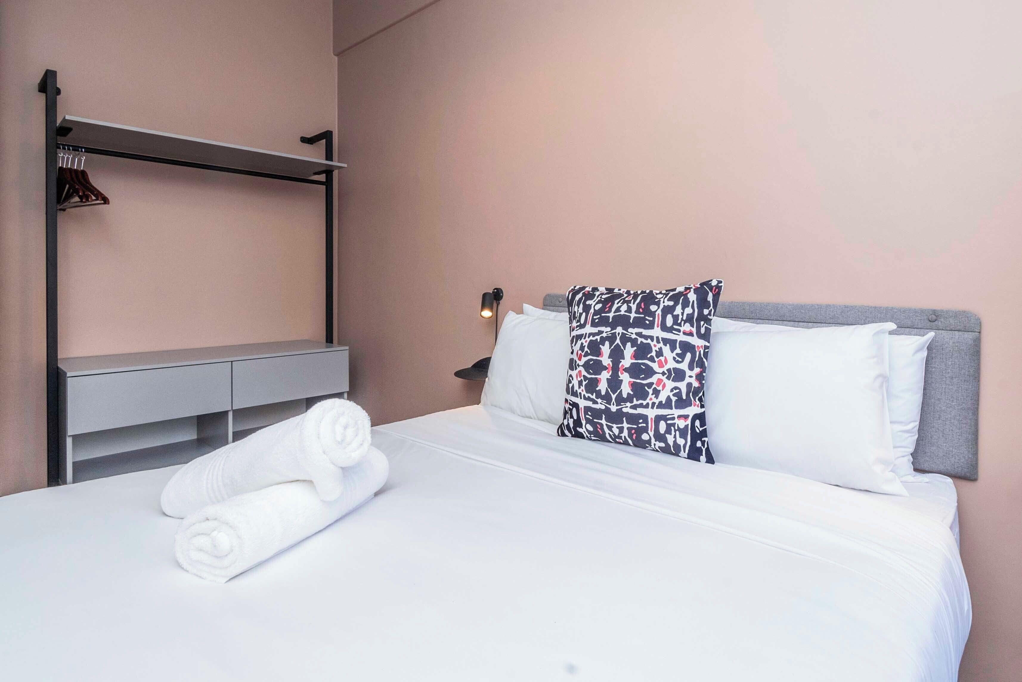 A hotel room with a bed, white linens, a towel, a headboard with patterned pillows, a bedside table, and a wall-mounted shelf with hangers.