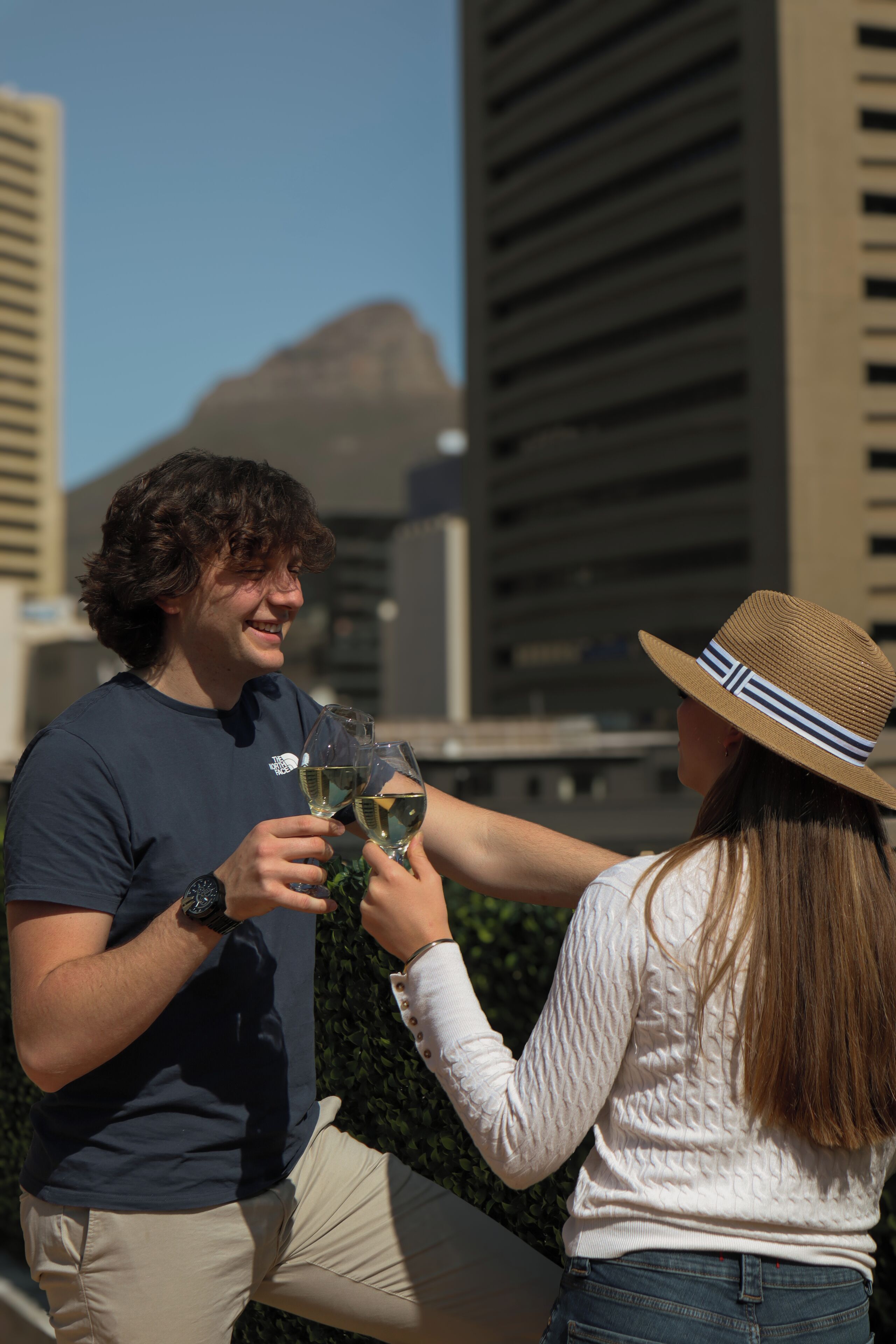 Una persona sosteniendo una copa de vino, otra persona con sombrero, y un paisaje urbano con edificios al fondo.