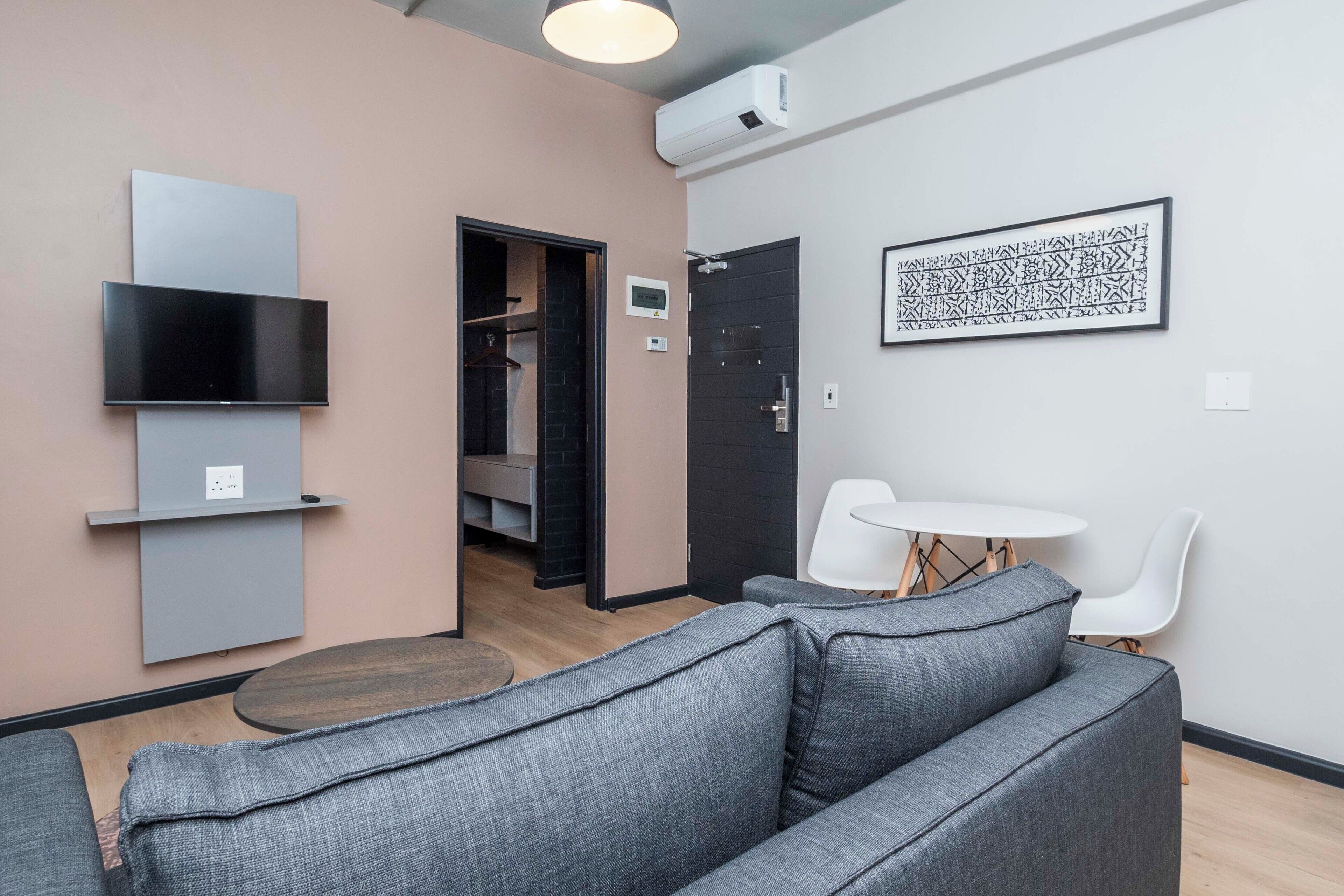 A modern living room with a grey sofa, a small round table, and a white chair. There is a TV mounted on the wall and a framed artwork above the door.