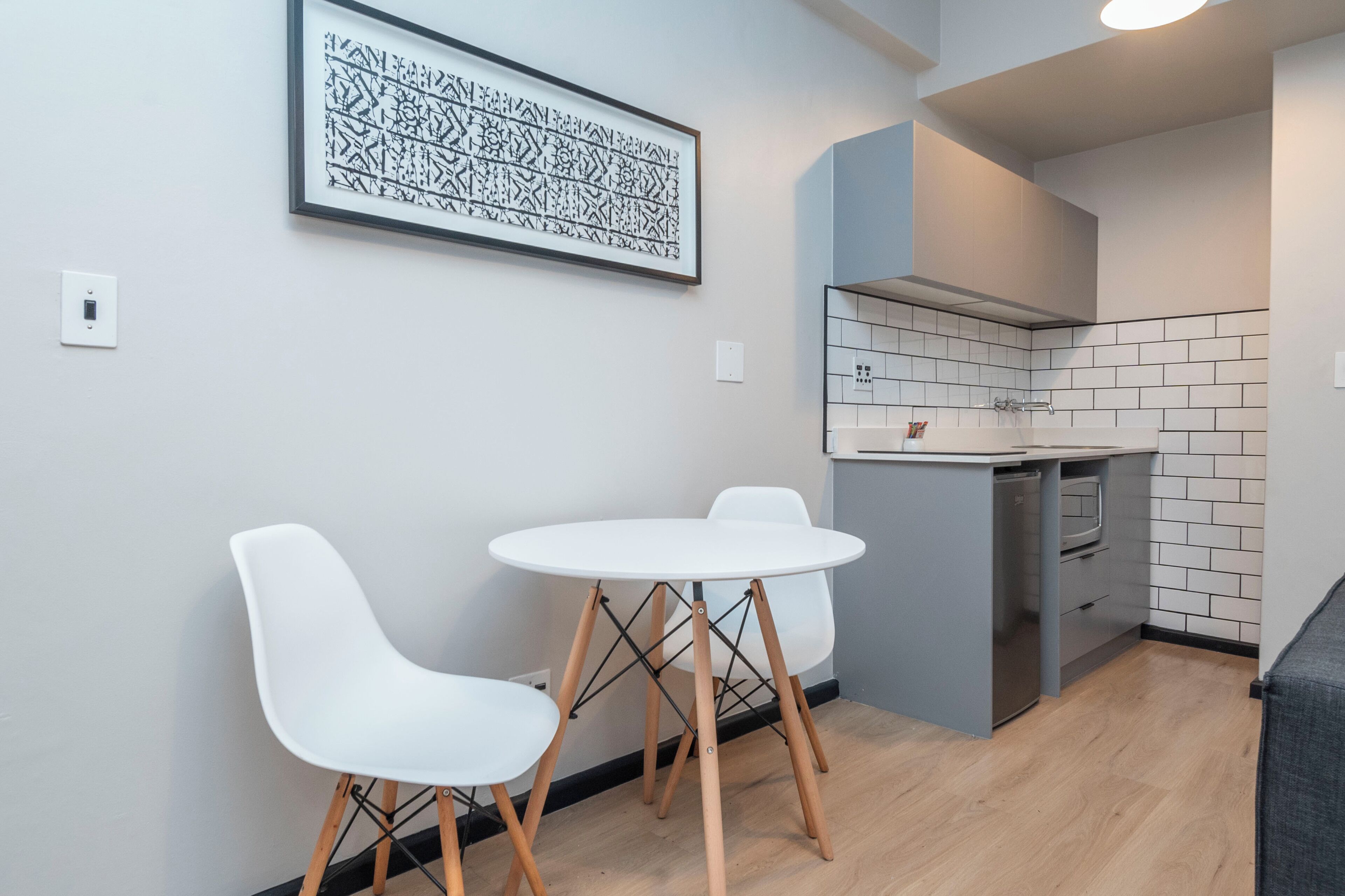 Una cocina moderna con armarios blancos, horno empotrado y encimera gris. Hay una mesa de comedor blanca con dos sillas.