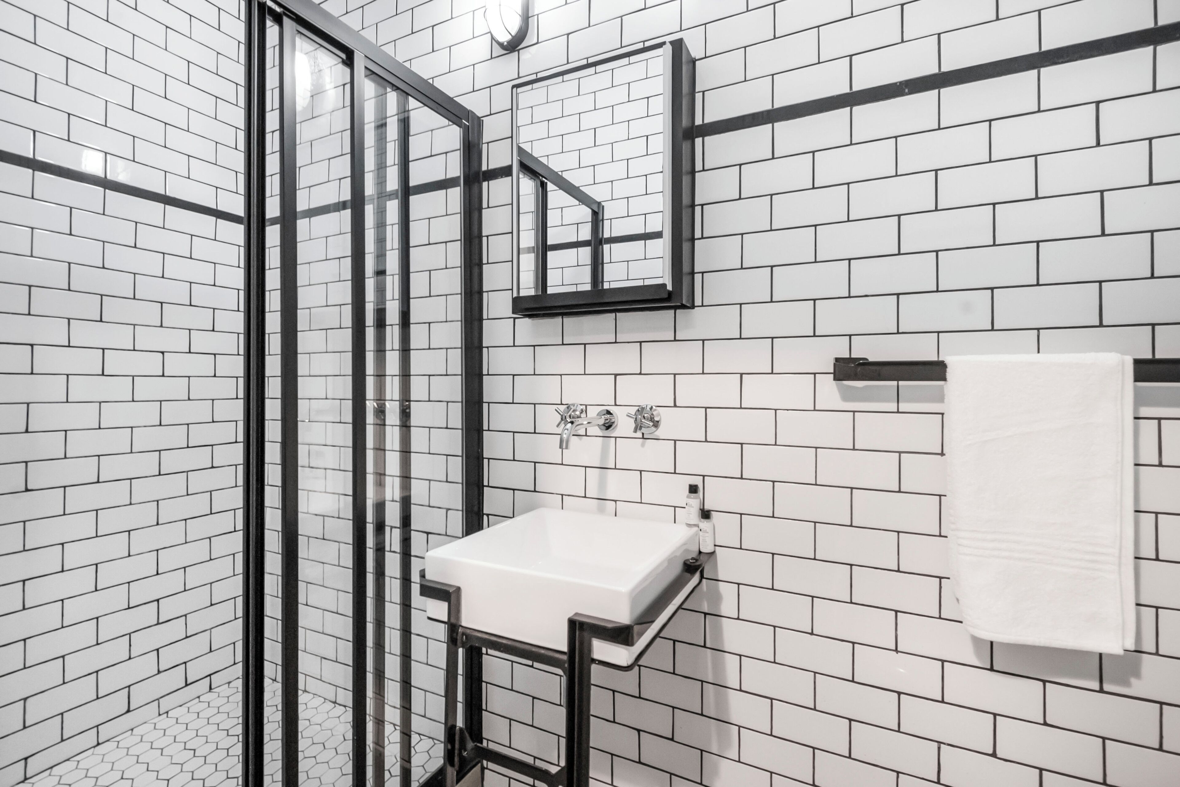 Une salle de bain avec des carreaux de céramique blancs rectangulaires, une douche en verre, un lavabo blanc et un porte-serviettes.