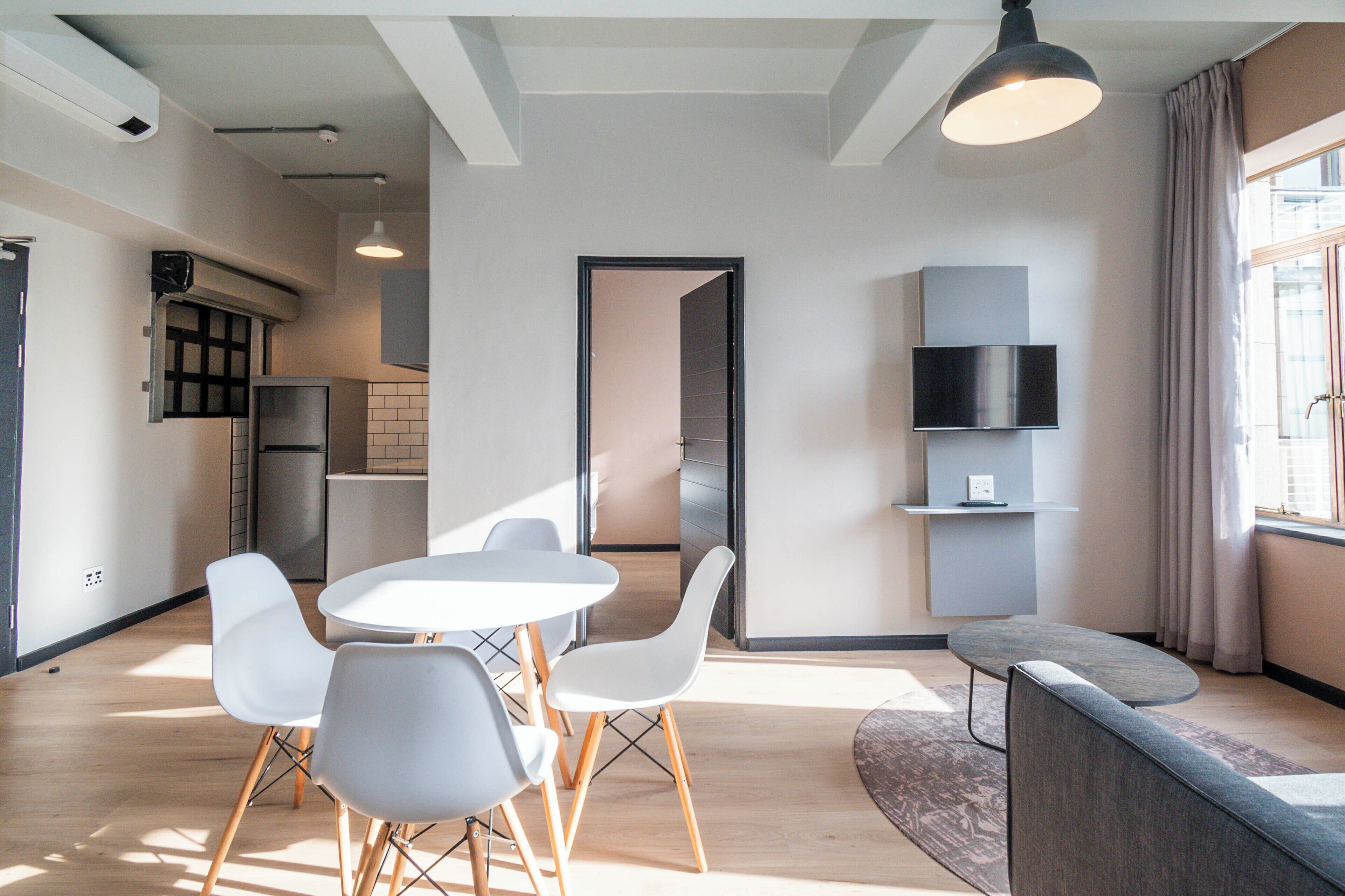 A modern kitchen with white cabinets, a built-in oven, and a refrigerator. There is a dining area with a round table and four chairs. A flat-screen TV is mounted on the wall.