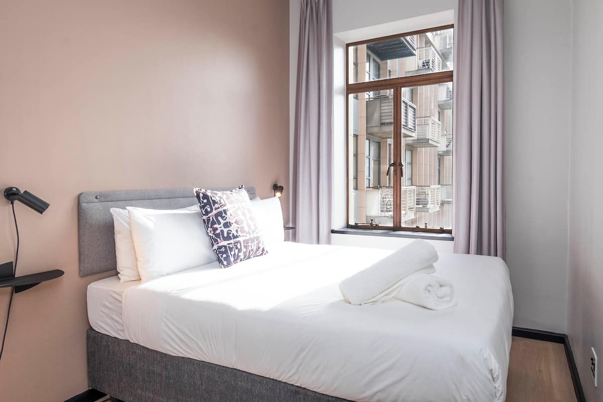 Una cama bien hecha con sábanas blancas y un cojín estampado, una lámpara de noche y una ventana con cortinas.
