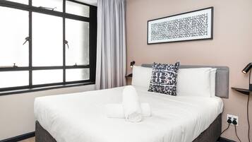 Una cama bien hecha con sábanas blancas, un cabecero gris y una almohada estampada. Un cuadro enmarcado cuelga de la pared sobre la cama.