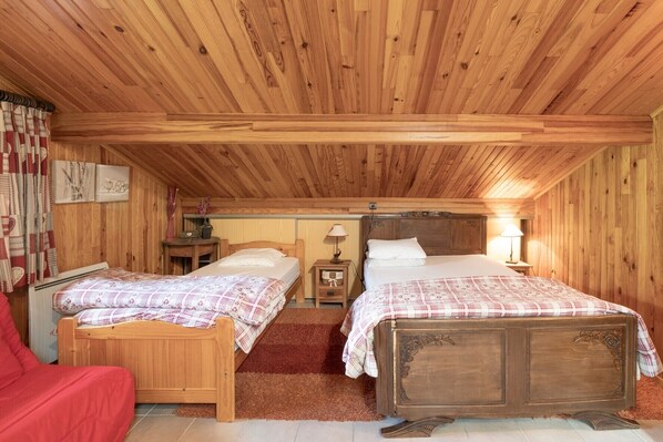 A wooden cabin bedroom with two beds, a red chair, and a small table with a lamp.