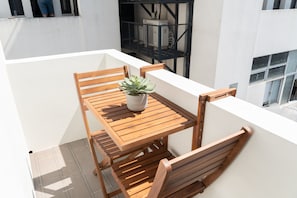 A wooden table with chairs and a potted plant on a balcony with white walls and a black railing.