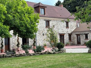 A stone house with a tiled roof, multiple windows, and a well-maintained garden with potted plants and a lawn. - Le Magnolia (Épieds)
