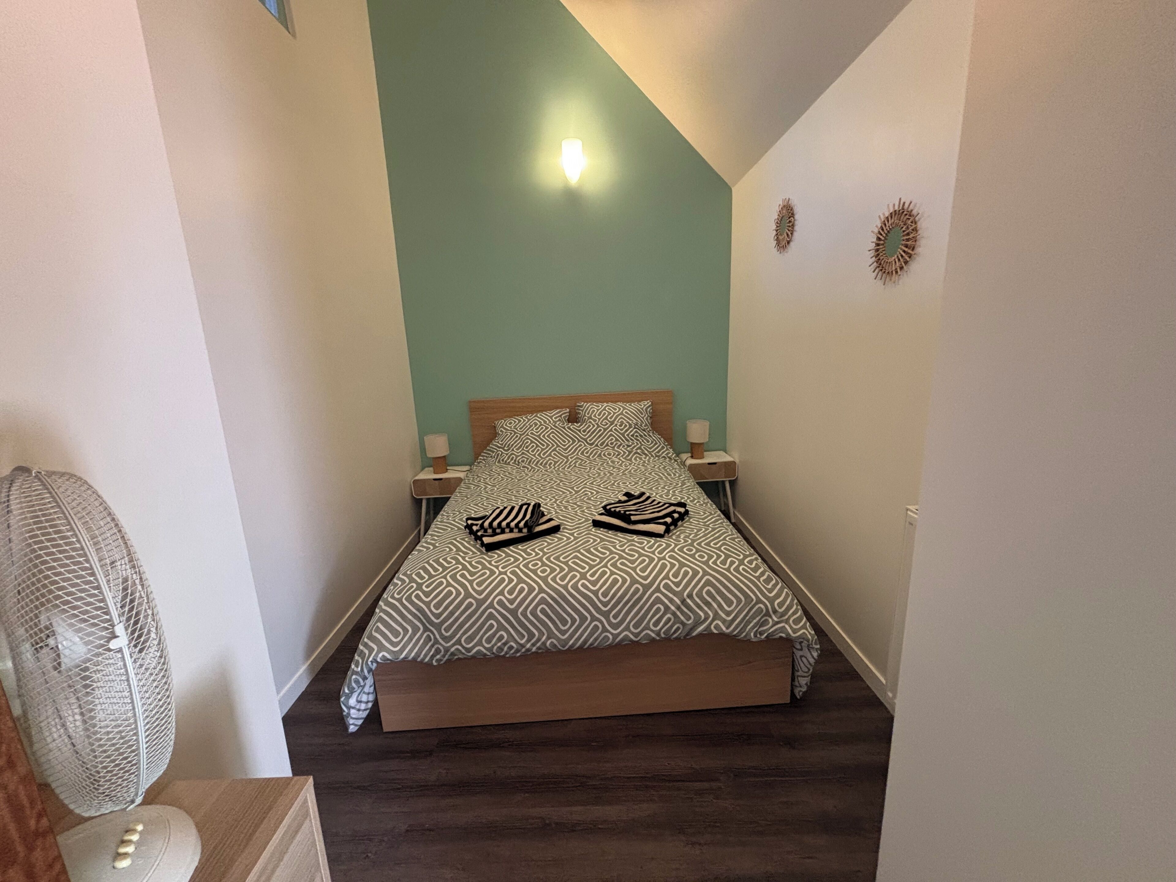 A bedroom with a bed, two bedside tables, a wall-mounted light, and a standing fan.