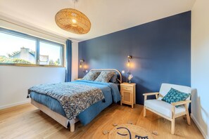 A bedroom with a bed, nightstand, chair, and a large window.