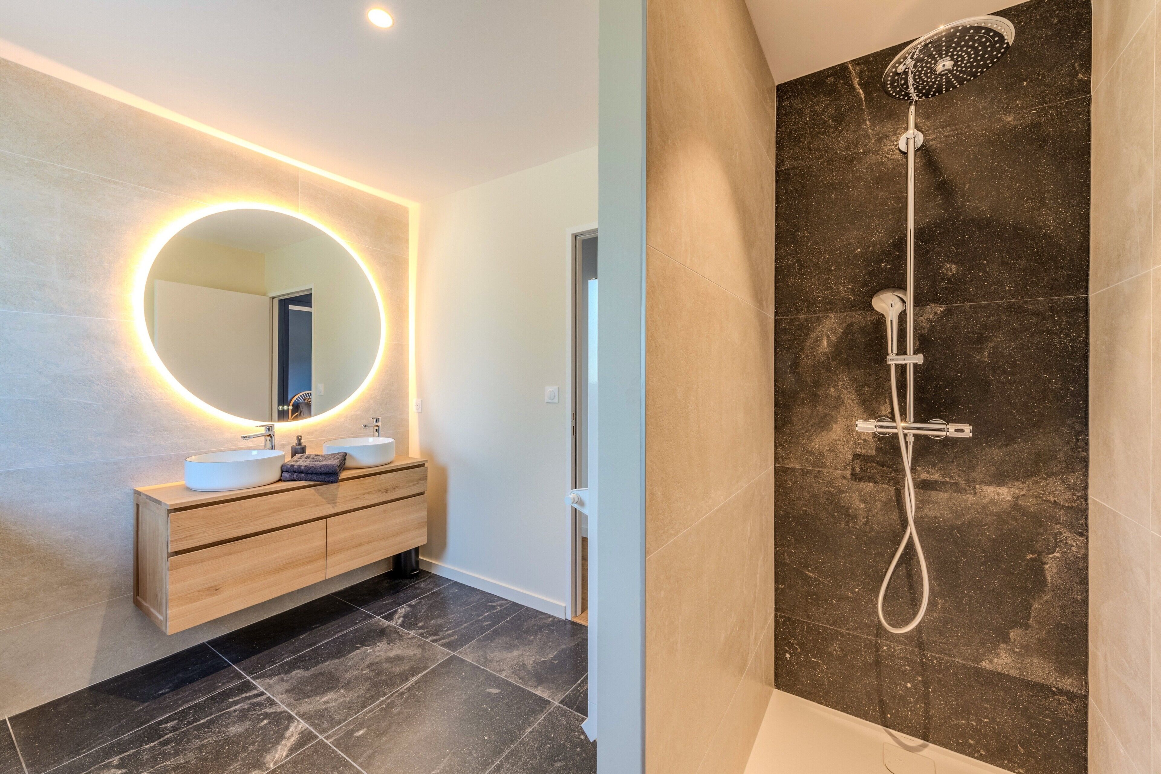 A modern bathroom with a round mirror, two sinks, a wooden vanity, and a shower with a handheld showerhead.
