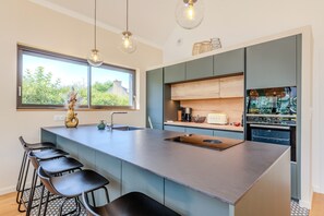 A modern kitchen with a large island, bar stools, a built-in oven, and a window with a view of greenery.