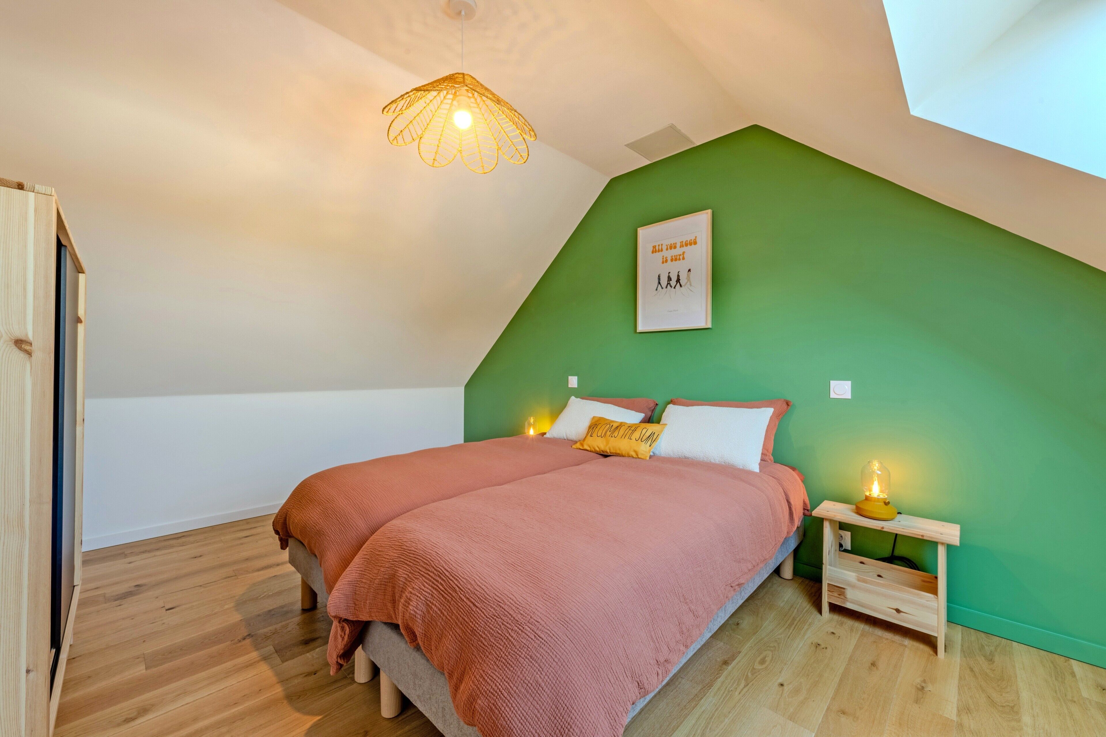 A bedroom with a sloped ceiling, a bed with pink bedding, a green accent wall, a small bedside table with a lamp, and a framed quote on the wall.