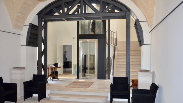 A spacious interior with a high arched doorway leading to a staircase. There are two black armchairs and a small table with a bowl on a pedestal. - Palazzo della Torre 003 - Historical modern stay (Bari)