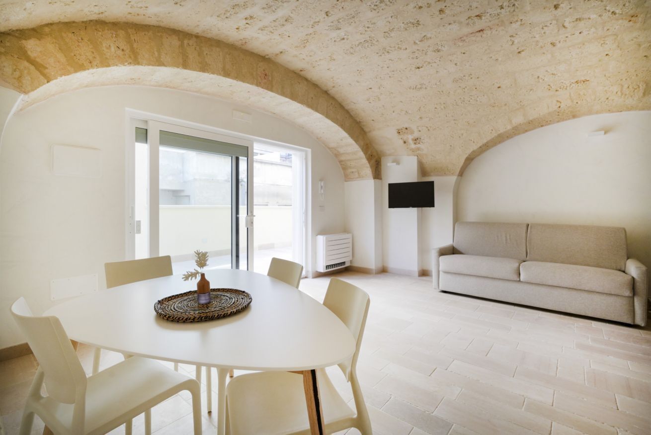 Zona pranzo con tavolo e sedie bianche, soffitto con mattoni a vista, divano e televisore a schermo piatto montato a parete.