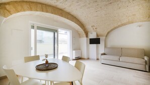 A dining area with a white table and chairs, a ceiling with exposed brickwork, a sofa, and a flat-screen TV mounted on the wall. - Palazzo della Torre 003 - Historical modern stay (Bari)