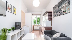 A modern living room with a grey sofa, a white coffee table, and a TV on a white stand. There are two framed pictures on the wall, a kitchen with a window, and a large painting on the wall. - ShortStayPoland Józefitów C4 (Kraków)