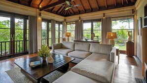 A spacious living room with a large sectional sofa, a coffee table, and a ceiling fan. Large windows provide a view of the outdoors. - Specious  three bedrooms villa in beautiful Jimbaran with private pool (Bali)