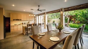 A dining area with a wooden table set for four, surrounded by chairs, in a modern kitchen with light-colored cabinets and a view of a lush garden. - Specious  three bedrooms villa in beautiful Jimbaran with private pool (Bali)