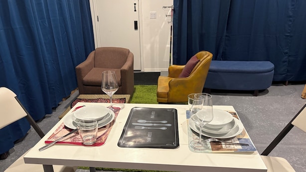 Une table à manger dressée avec des assiettes, des verres et des couverts, entourée de chaises et d’un canapé, dans une pièce avec des rideaux bleus et une porte.