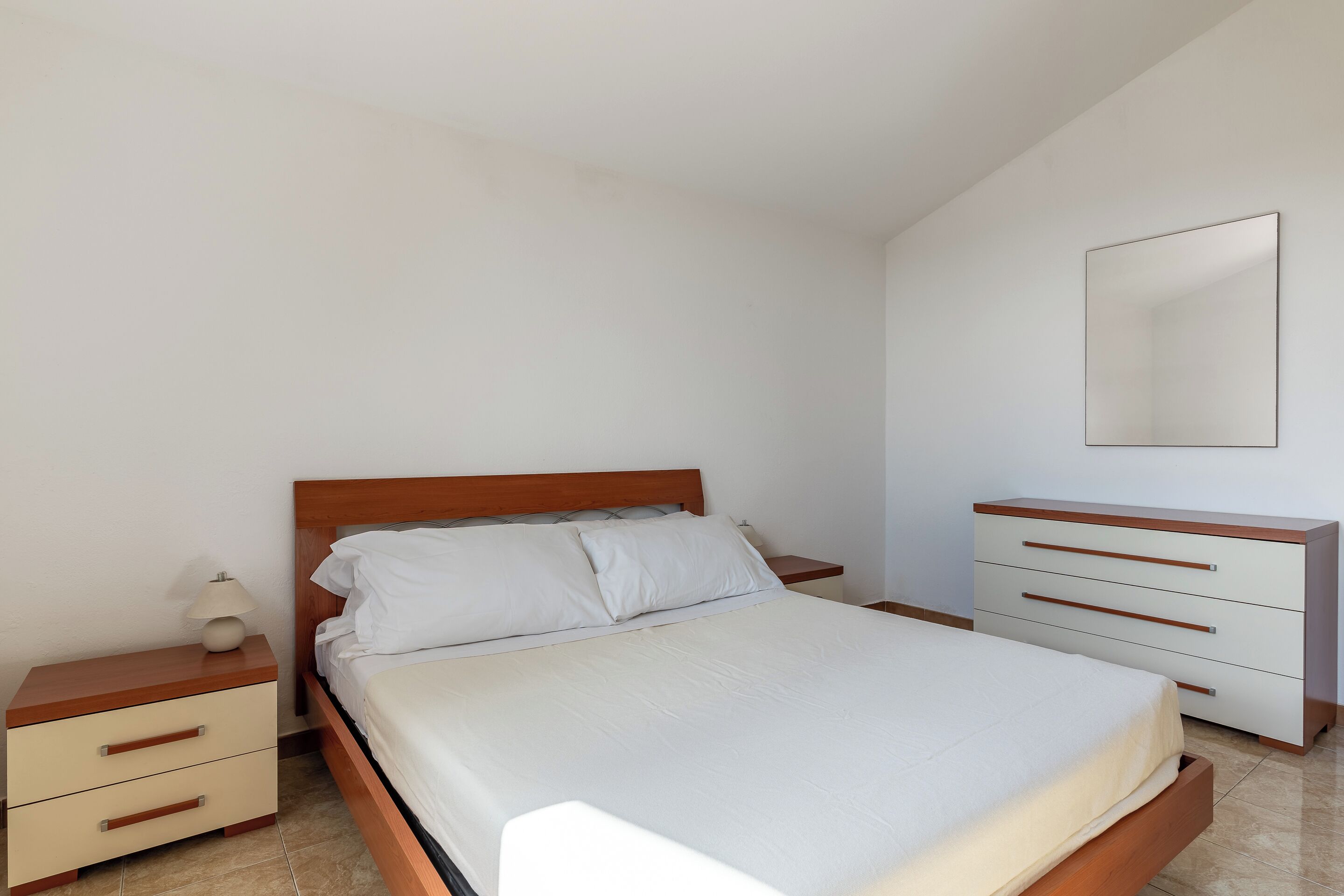 A bedroom with a wooden bed, a nightstand with drawers, a bedside table with a lamp, and a dresser with drawers.