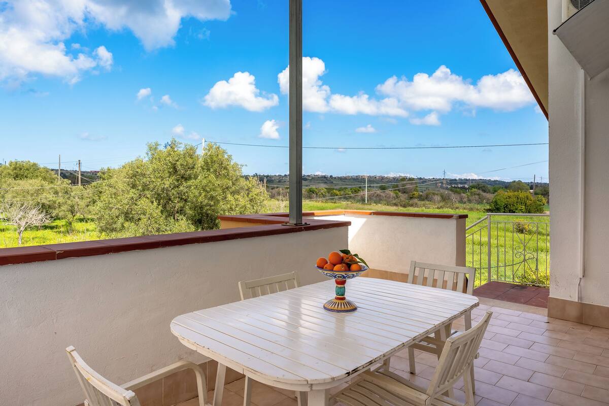 A balcony with a white table and chairs, a bowl of fruit, and a view of a green landscape.