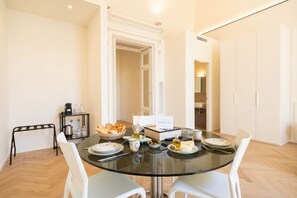 A dining area with a glass table, white chairs, a coffee machine, and a buffet with bread and cheese. - Palazzo della Torre 102 - Luxury modern suite (Bari)