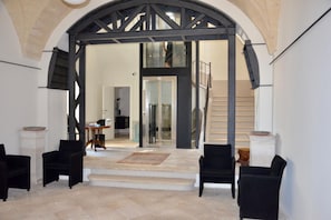 A spacious interior with a high arched doorway leading to a staircase. There are two black armchairs and a small table with a bowl on a pedestal.