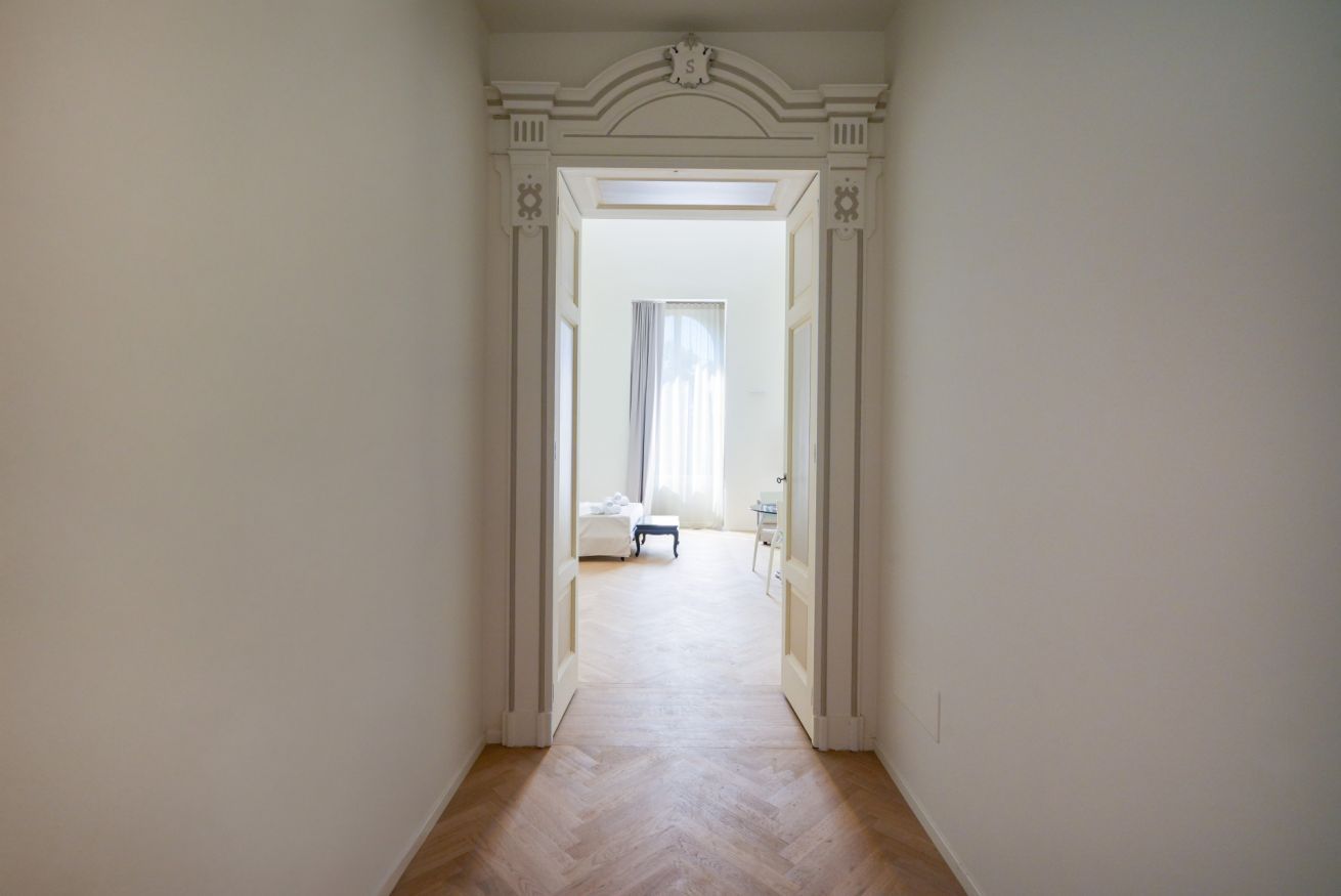 Un corridoio con una porta bianca che dà su una stanza con un letto e una sedia.