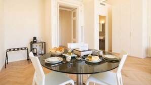 Un comedor con mesa de vidrio, sillas blancas, una máquina de café y un bufé con pan y queso.
