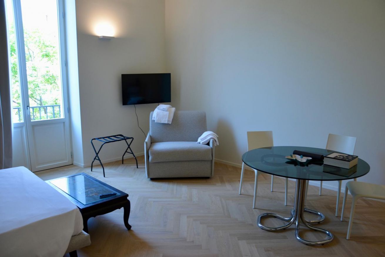 Hotelkamer met een bed, een glazen salontafel, een grijze fauteuil, een ronde glazen tafel met stoelen, een aan de muur bevestigde televisie en een raam met uitzicht op bomen.
