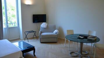 Hotelkamer met een bed, een glazen salontafel, een grijze fauteuil, een ronde glazen tafel met stoelen, een aan de muur bevestigde televisie en een raam met uitzicht op bomen.