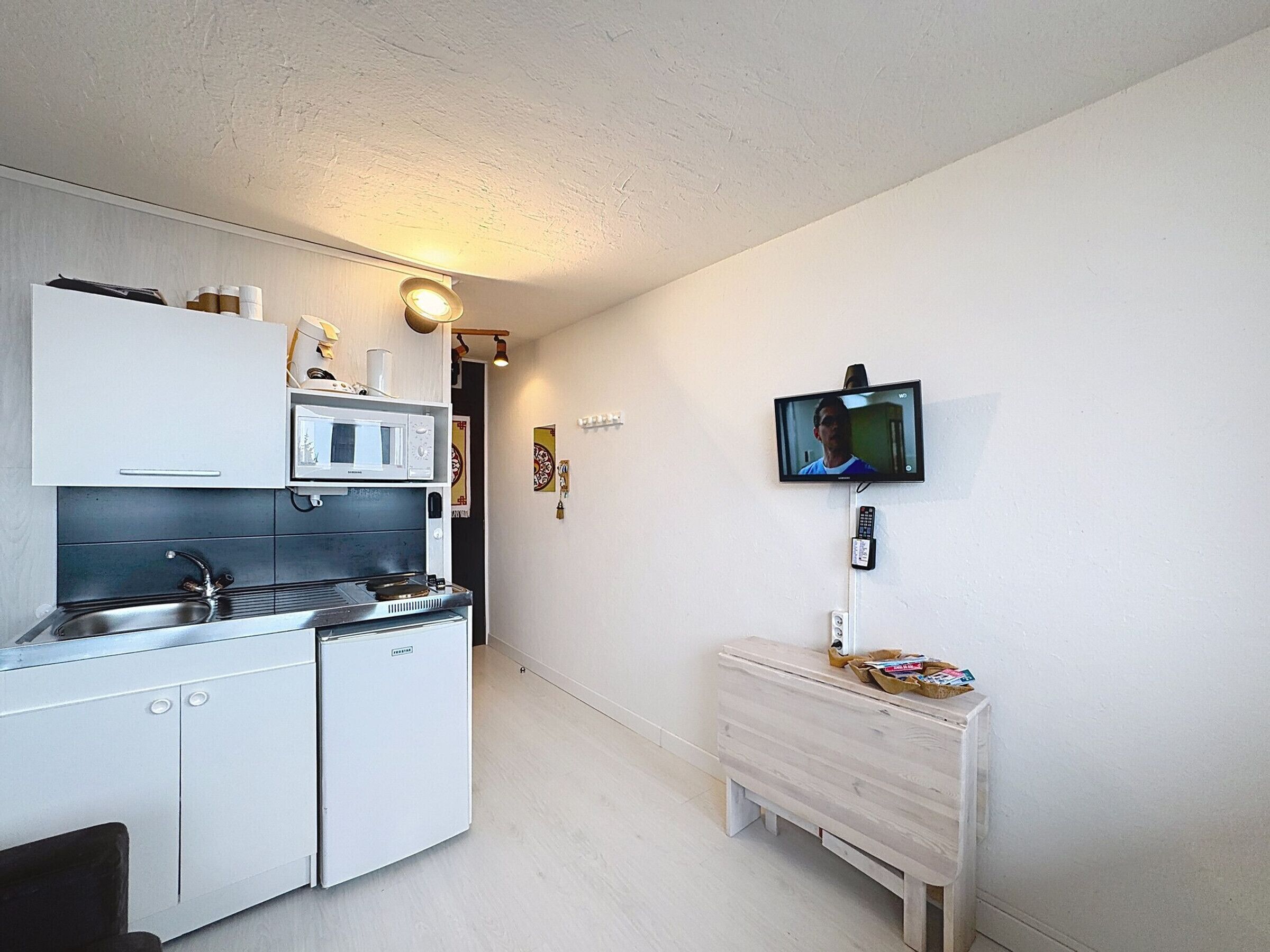 A compact kitchen with a microwave, stove, and sink. A TV is mounted on the wall, and a small wooden cabinet is against the wall.