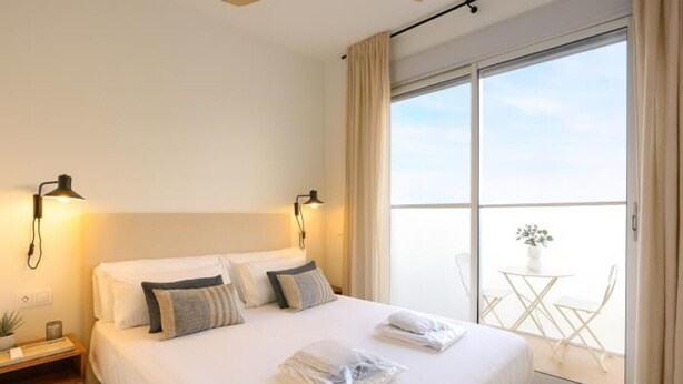A modern hotel room with a large bed, two bedside lamps, a small table with a plant, and a balcony with a table and chairs. - Modern apartment 5 min from the beach (Puerto de Sagunto)