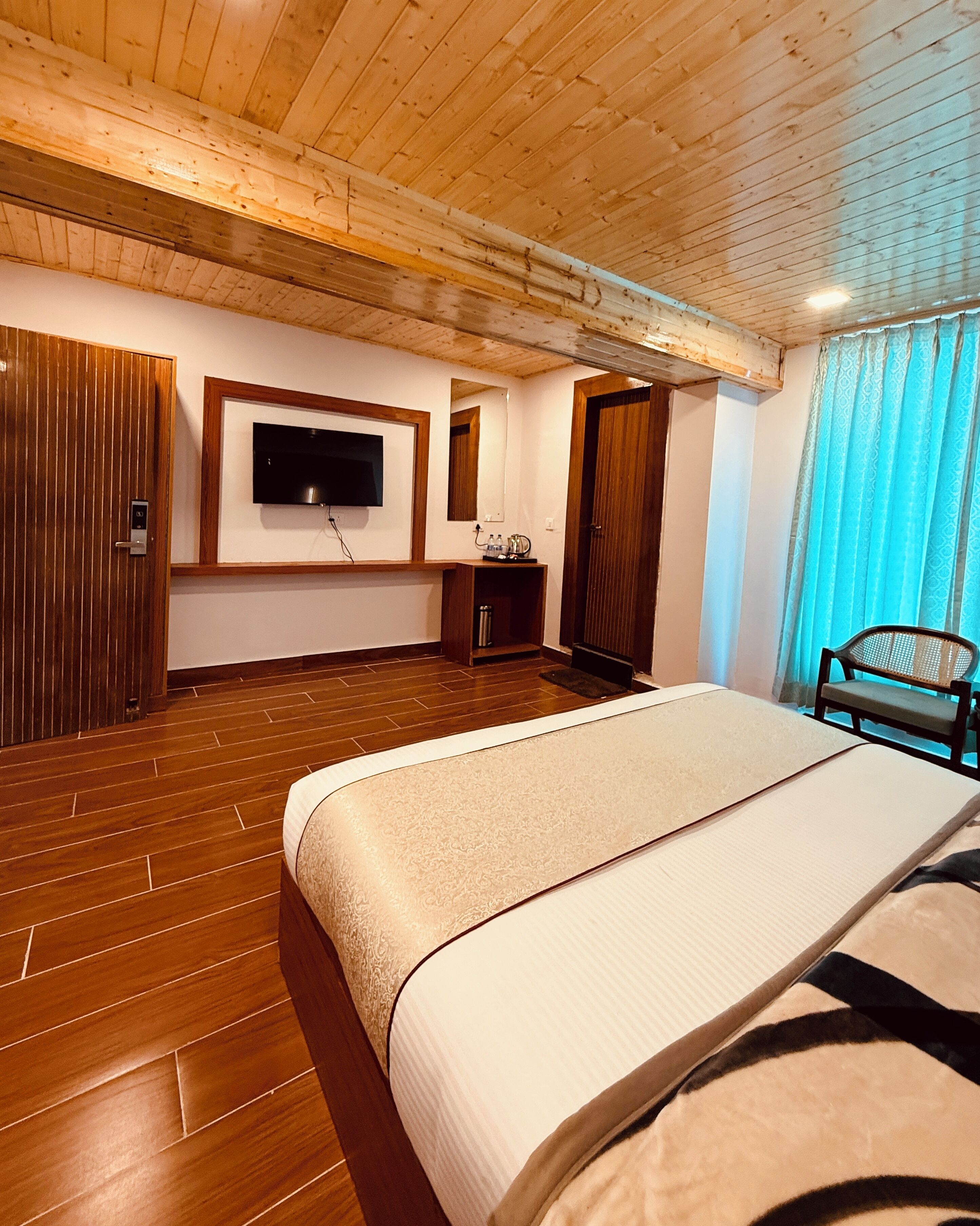 Une chambre d’hôtel avec un plancher en bois, un lit recouvert d’une couvre-lit beige, un téléviseur fixé au mur, une chaise et une fenêtre avec des rideaux bleus.