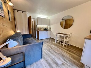 A compact living space with a sofa, a dining table with chairs, a kitchen area, and a round mirror on the wall.
