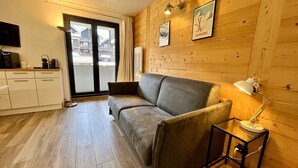 A cozy room with a sofa, a small table with a lamp, a kitchen area, and wooden walls. - Renovated studio in village center with balcony (Samoëns)