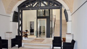 Un interior espacioso con un arco alto que da a una escalera. Hay dos sillones negros y una pequeña mesa con un cuenco sobre un pedestal.