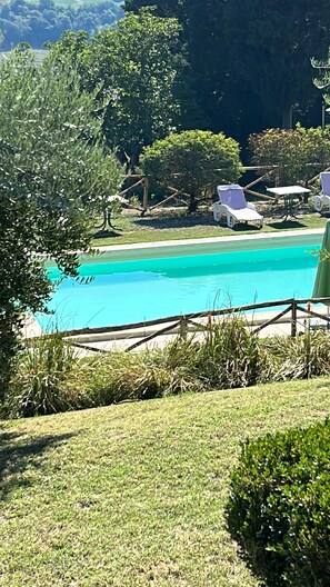 Ein Pool mit klarem, blauem Wasser, umgeben von einer Rasenfläche und Bäumen. Liegestühle und ein Tisch befinden sich in Poolnähe.