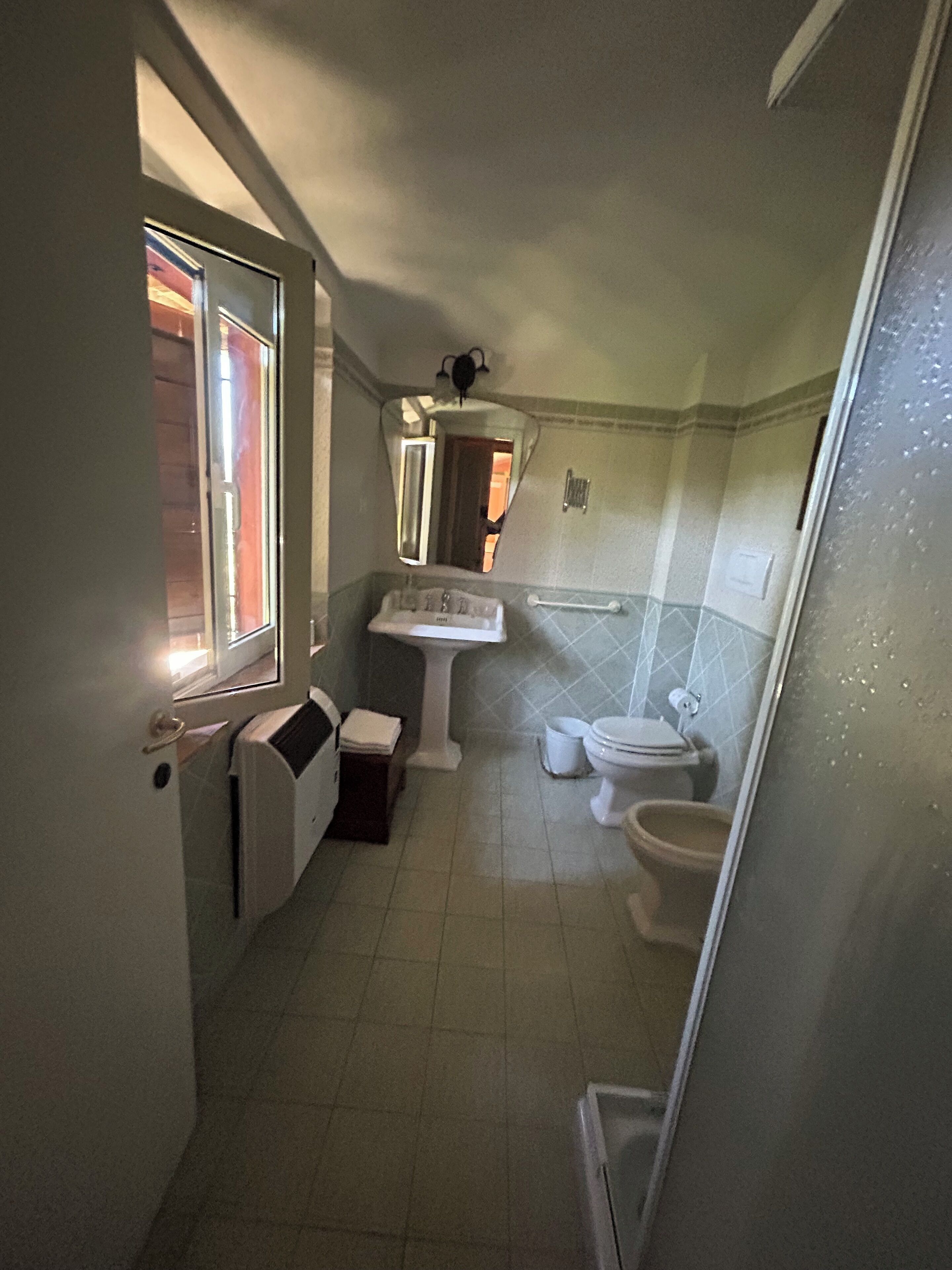 A small, compact bathroom with a window, a pedestal sink, a toilet, and a radiator.