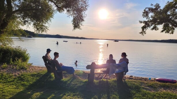 A lakeside setting with people relaxing on a bench, a boat on the water, and a clear view of the sunset.