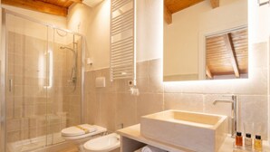 A bathroom with a glass-enclosed shower, a toilet, a sink with a mirror, and towels on a shelf.