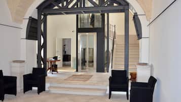 Un ampio interno con un'imponente porta ad arco che conduce a una scala. Si notano due poltrone nere e un tavolino con una ciotola su un piedistallo.