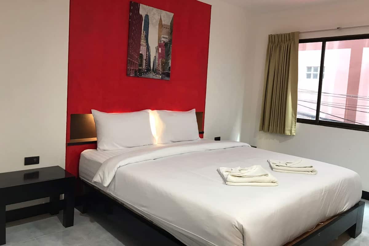 A hotel room with a bed, a nightstand, a red accent wall, a window with curtains, and a cityscape painting.