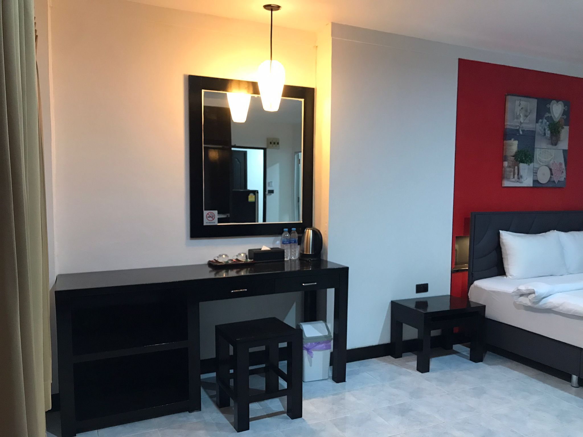 A hotel room with a black desk, a small stool, a bed with white bedding, a red accent wall, and a large mirror with a light fixture above it.