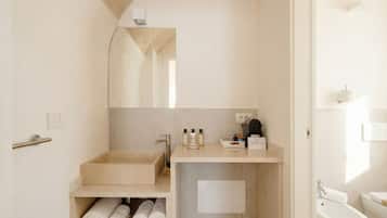 Ein modernes Badezimmer mit einem beigen Waschtisch, einem Spiegel und einer Toilette. Auf der Ablage des Waschtisches liegen Handtücher und Pflegeprodukte.